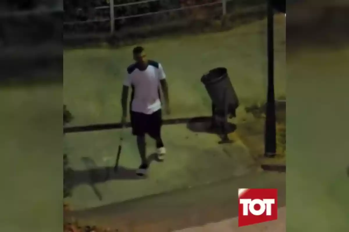 Un hombre con camiseta blanca y pantalones cortos oscuros camina por la acera de noche sosteniendo un objeto largo cerca de un bote de basura y un poste de luz