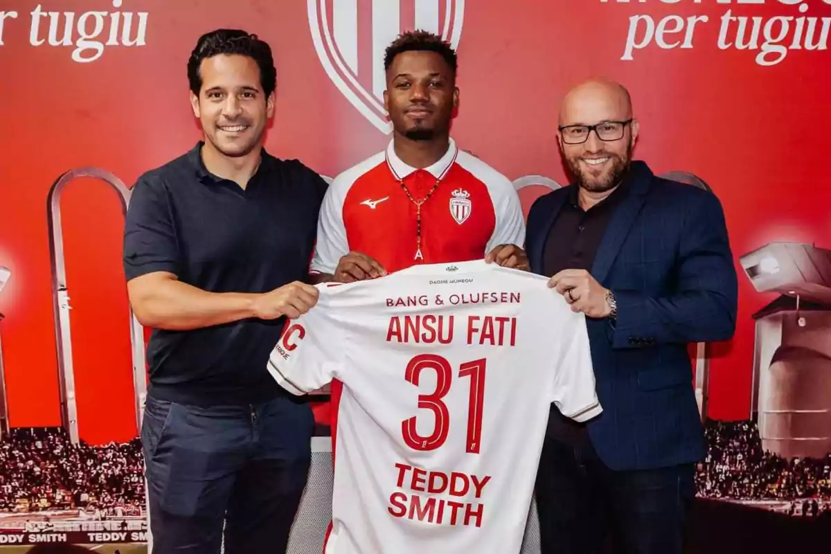 Tres personas posan sonrientes mientras sostienen una camiseta de fútbol del Mónaco con el nombre Ansu Fati y el número 31 frente a un fondo rojo con el escudo del club. Tres personas posan sonrientes mientras sostienen una camiseta de fútbol del Mónaco con el nombre Ansu Fati y el número 31 frente a un fondo rojo con el escudo del club.
