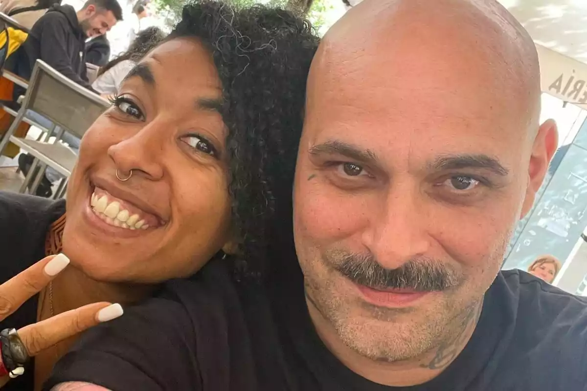 Dos personas sonríen y posan juntas para una selfie en un lugar al aire libre, una de ellas hace el signo de la paz con los dedos.