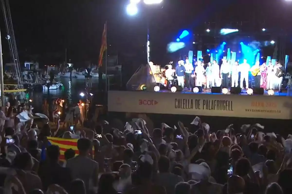 Un grupo de personas disfruta de un concierto nocturno al aire libre en Calella de Palafrugell con el público ondeando pañuelos blancos y una bandera catalana visible entre la multitud Un grupo de personas disfruta de un concierto nocturno al aire libre en Calella de Palafrugell con el público ondeando pañuelos blancos y una bandera catalana visible entre la multitud