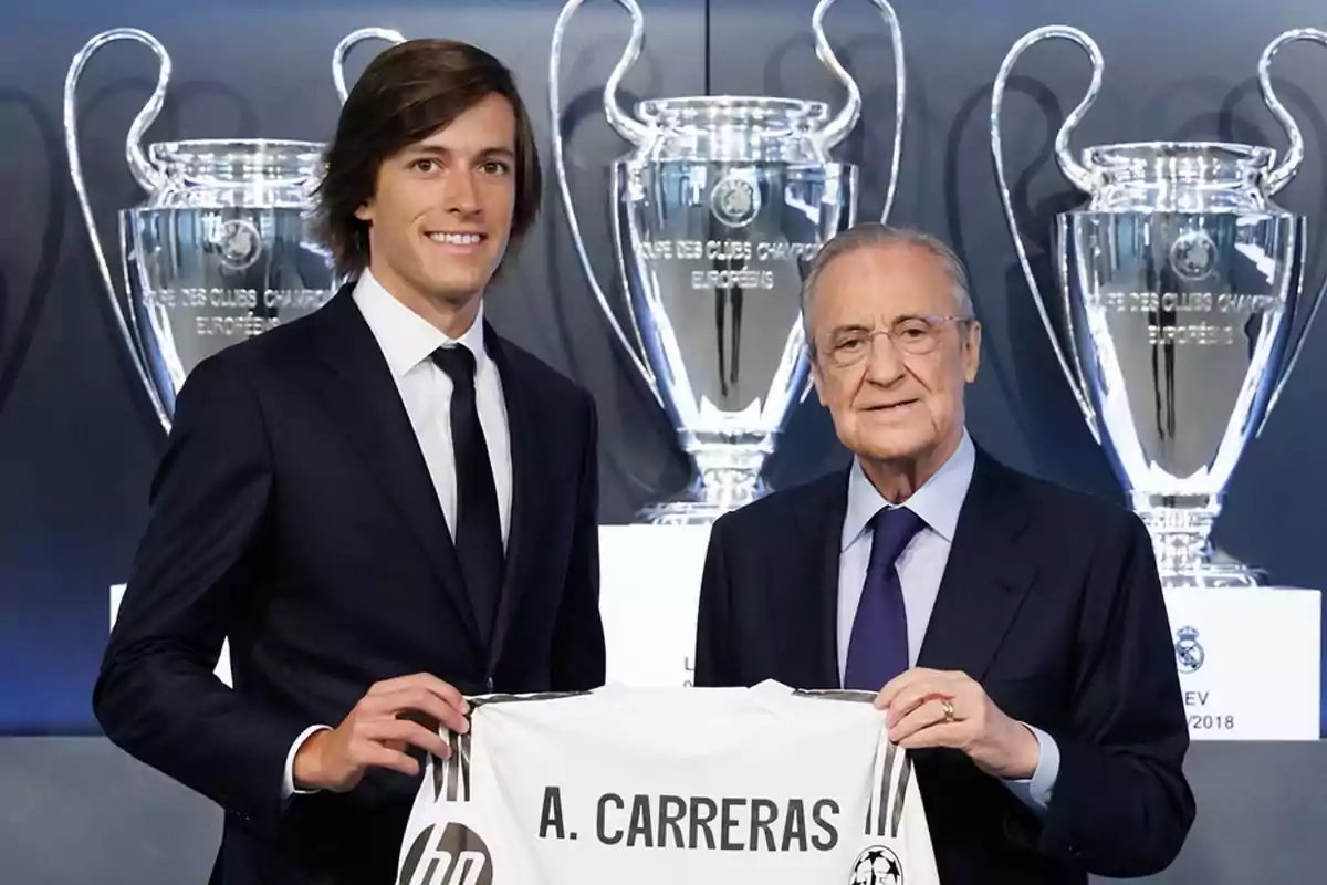 Dos hombres con traje posan sonrientes sosteniendo una camiseta de fútbol con el nombre A. Carreras frente a varios trofeos grandes de la Champions League. Dos hombres con traje posan sonrientes sosteniendo una camiseta de fútbol con el nombre A. Carreras frente a varios trofeos grandes de la Champions League.