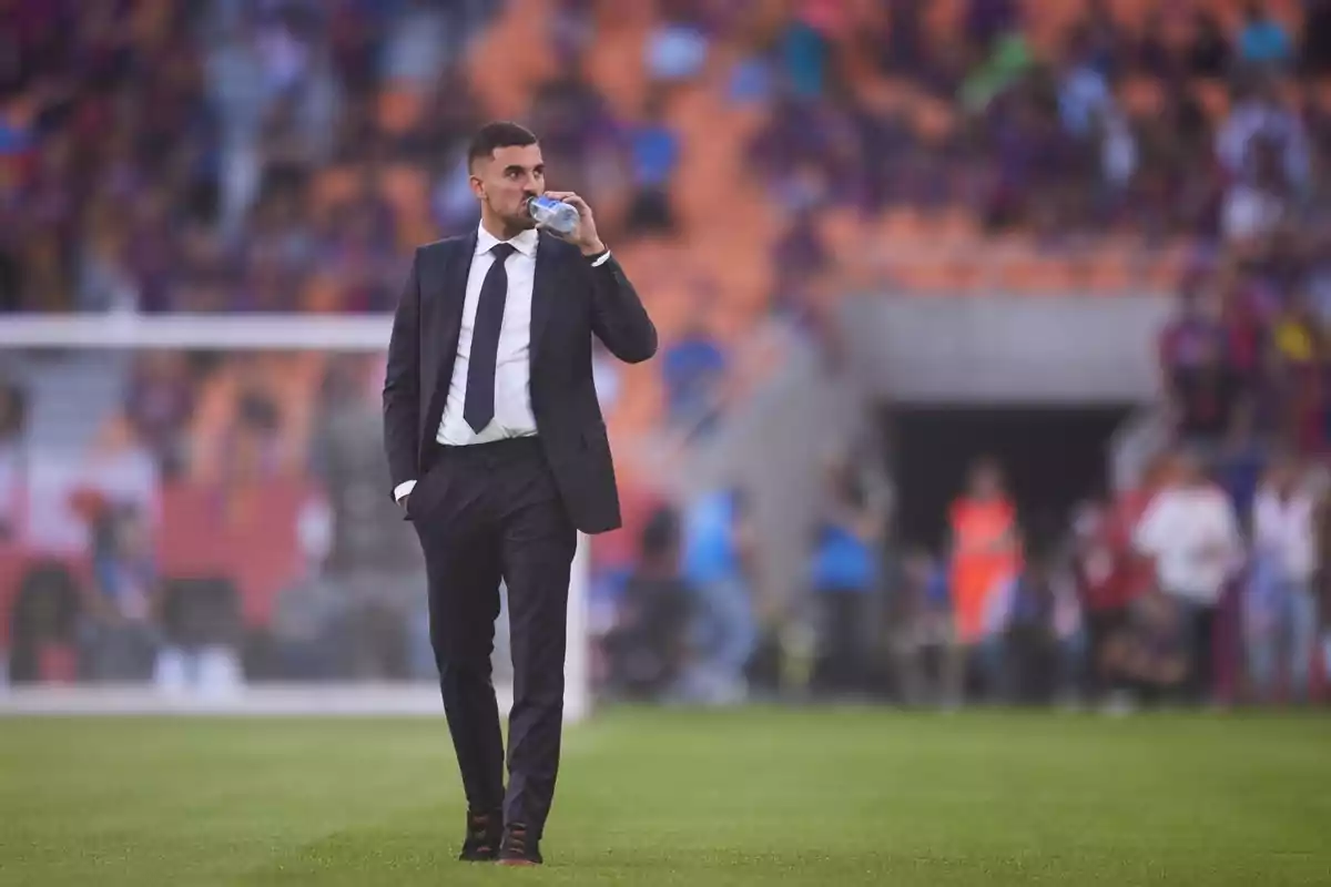 Hombre con traje oscuro bebiendo agua en el campo de fútbol durante un partido con público en las gradas al fondo Hombre con traje oscuro bebiendo agua en el campo de fútbol durante un partido con público en las gradas al fondo