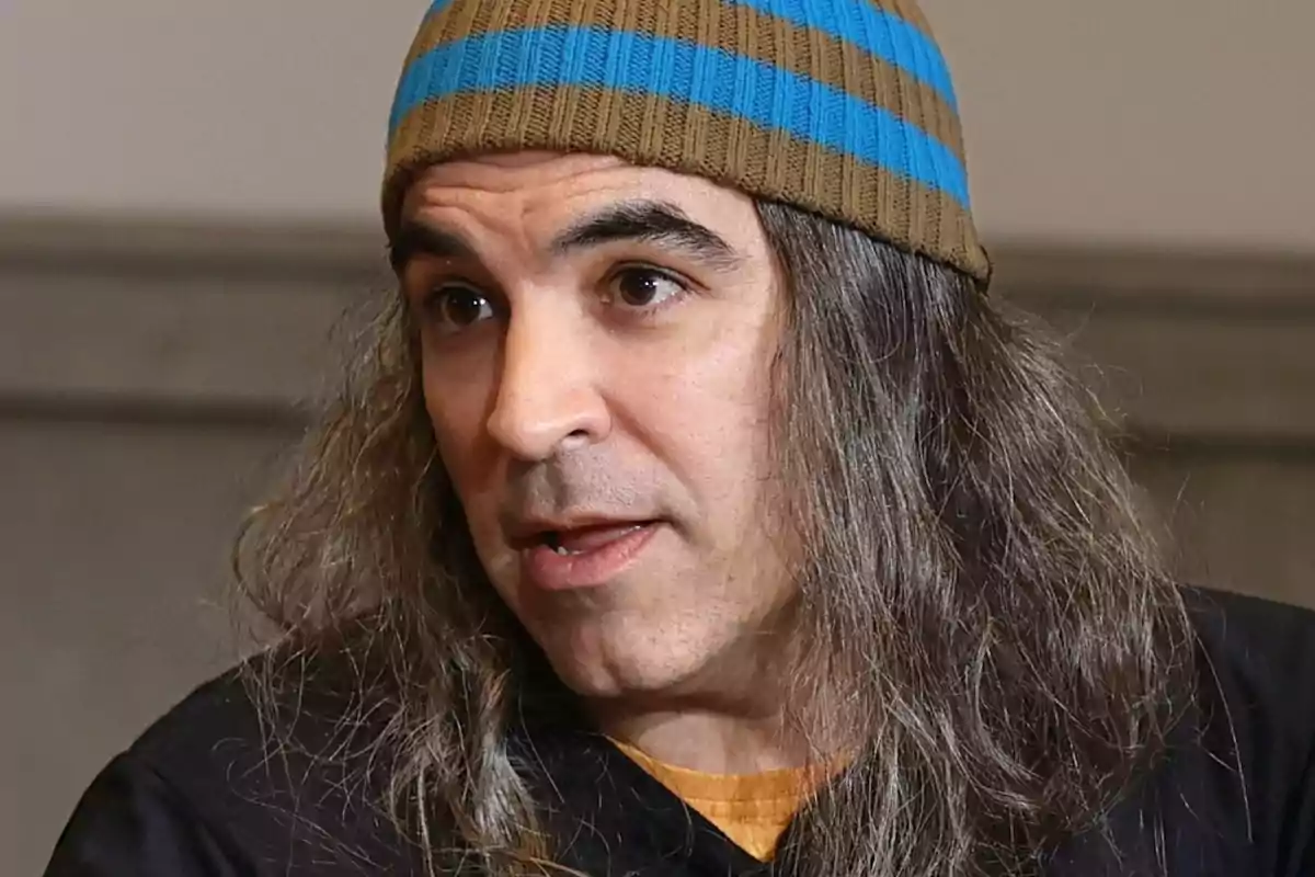 Hombre de cabello largo y canoso usando un gorro de rayas marrón y azul mientras habla