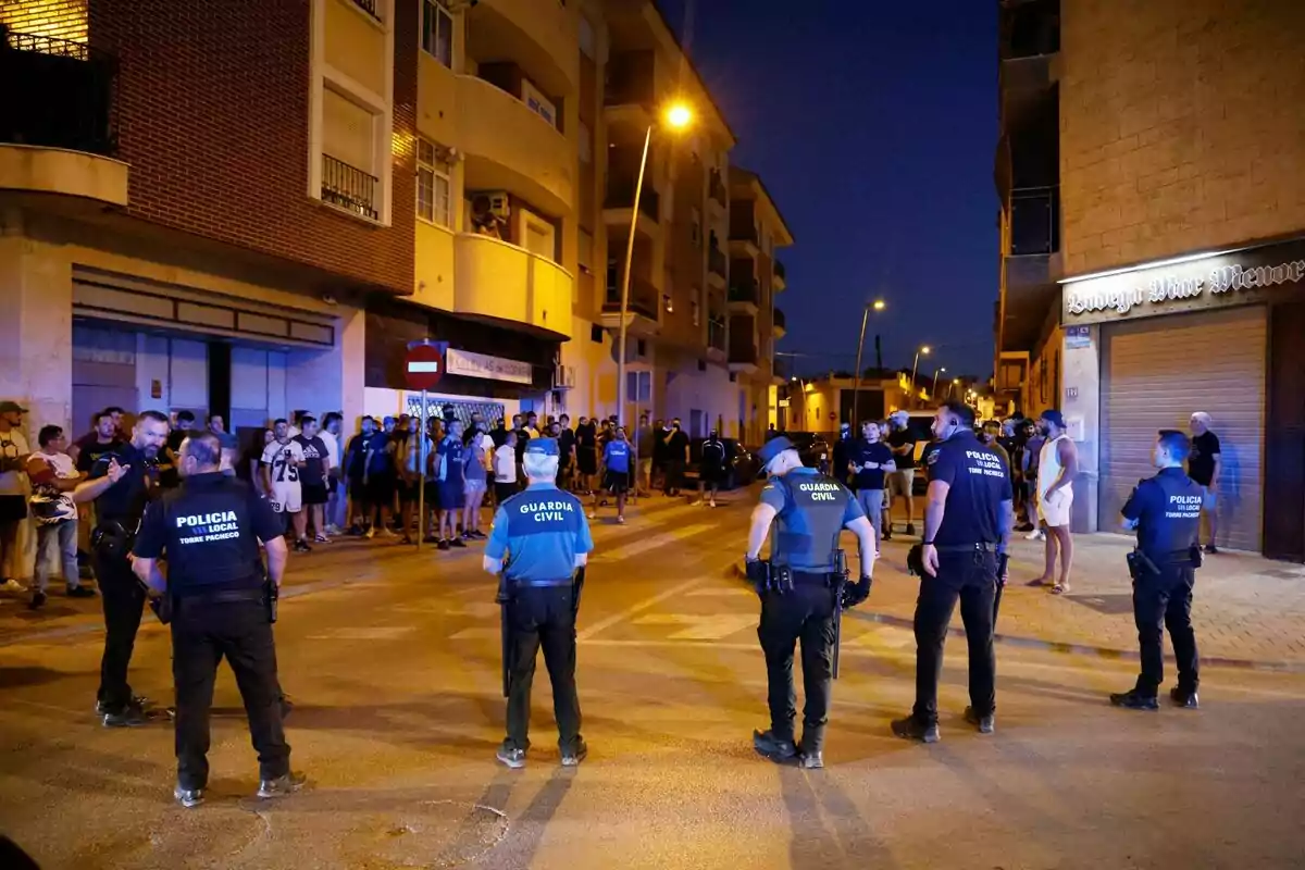 Varios policías forman una barrera en una calle durante la noche mientras un grupo de personas observa desde la acera frente a edificios residenciales iluminados por farolas. Varios policías forman una barrera en una calle durante la noche mientras un grupo de personas observa desde la acera frente a edificios residenciales iluminados por farolas.