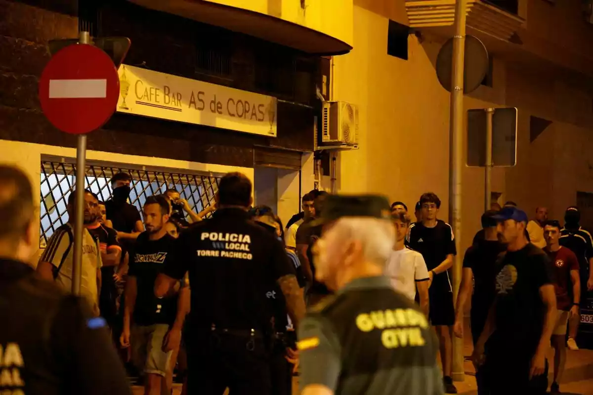 Un grupo de personas y agentes de policía se encuentran en la calle frente a un bar durante la noche Un grupo de personas y agentes de policía se encuentran en la calle frente a un bar durante la noche