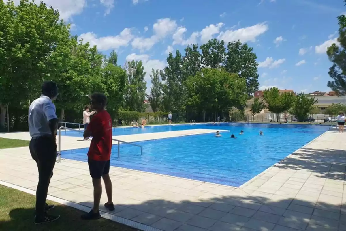 Personas conversando junto a una piscina al aire libre en un día soleado con varias personas nadando y árboles alrededor Personas conversando junto a una piscina al aire libre en un día soleado con varias personas nadando y árboles alrededor