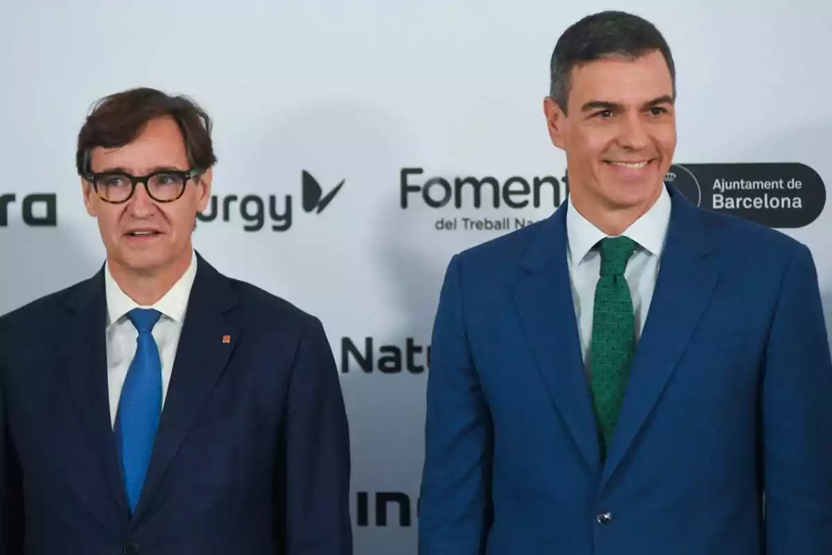 Dos hombres con traje posan frente a un fondo con logotipos de empresas y organizaciones