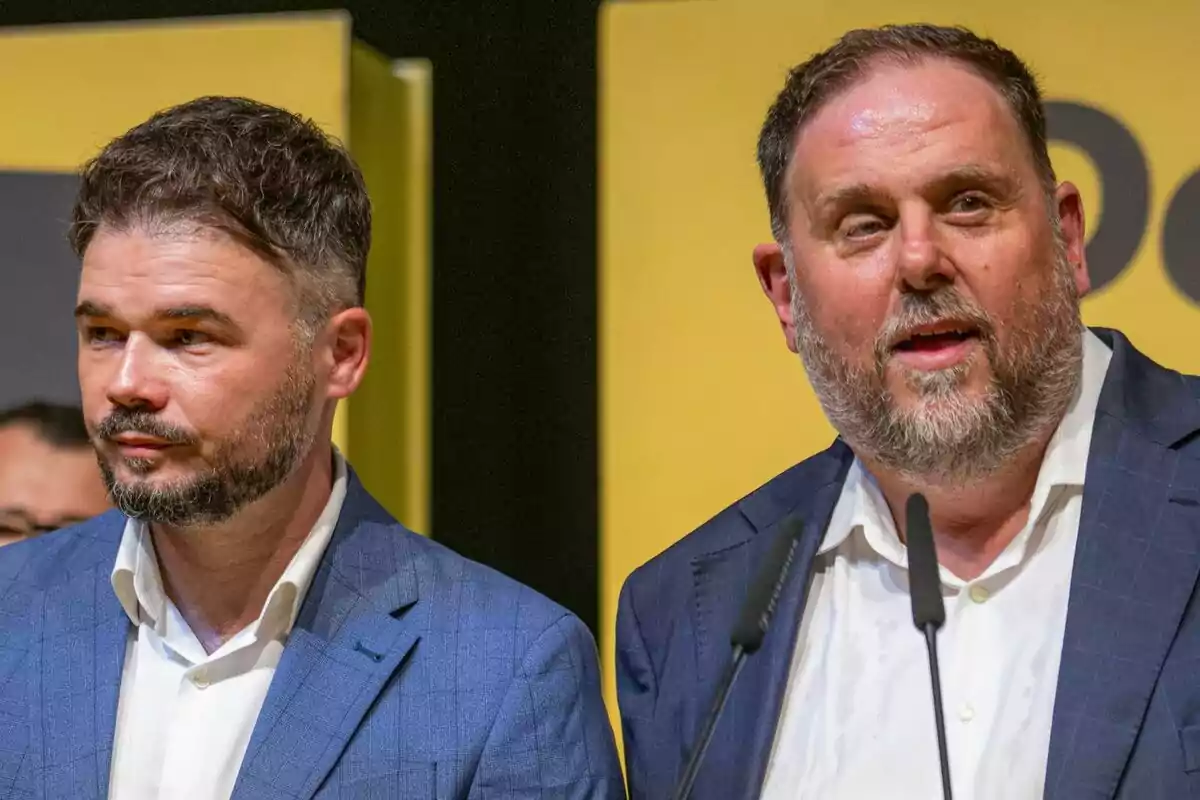 Dos hombres con barba y traje azul posan juntos frente a un fondo amarillo mientras uno de ellos habla frente a micrófonos