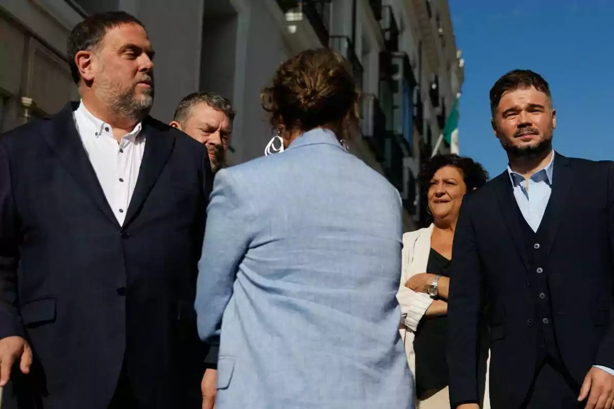 Cinco personas vestidas de manera formal conversan al aire libre en una calle con edificios y cielo despejado. Cinco personas vestidas de manera formal conversan al aire libre en una calle con edificios y cielo despejado.