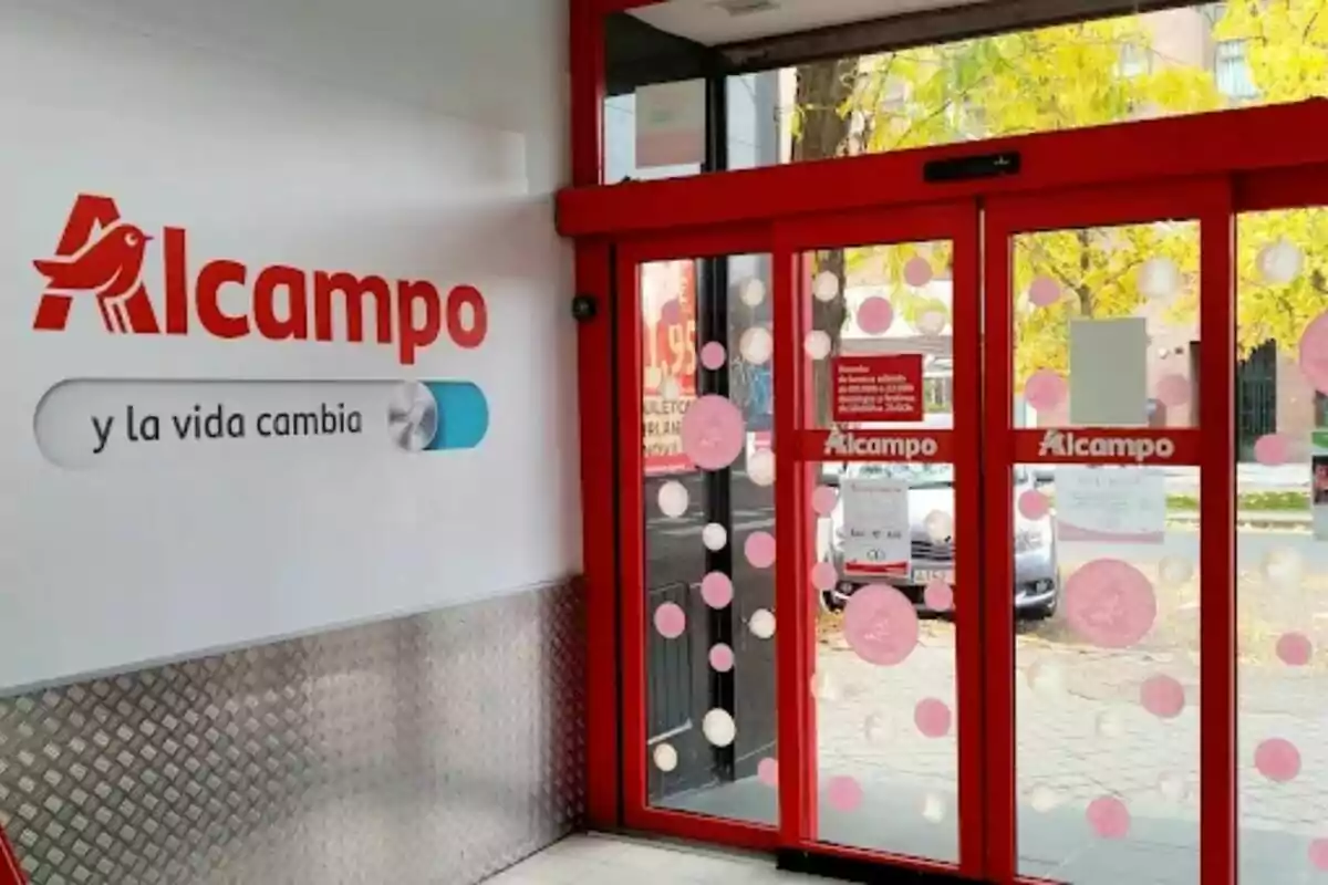 Entrada de supermercado Alcampo con puertas automáticas de cristal decoradas con círculos y el logo visible en la pared y en las puertas. Entrada de supermercado Alcampo con puertas automáticas de cristal decoradas con círculos y el logo visible en la pared y en las puertas.