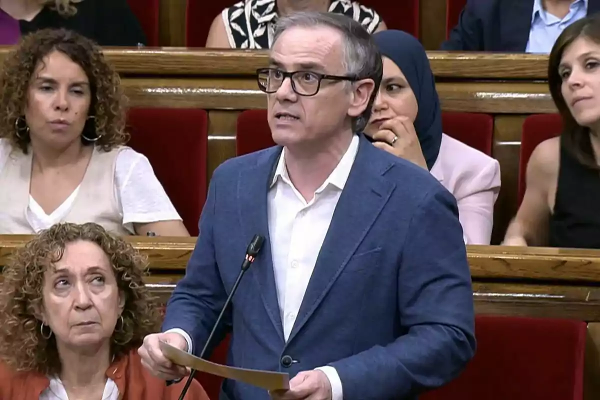 Hombre de gafas y chaqueta azul hablando en un parlamento mientras sostiene unos papeles rodeado de otras personas sentadas en escaños rojos Hombre de gafas y chaqueta azul hablando en un parlamento mientras sostiene unos papeles rodeado de otras personas sentadas en escaños rojos