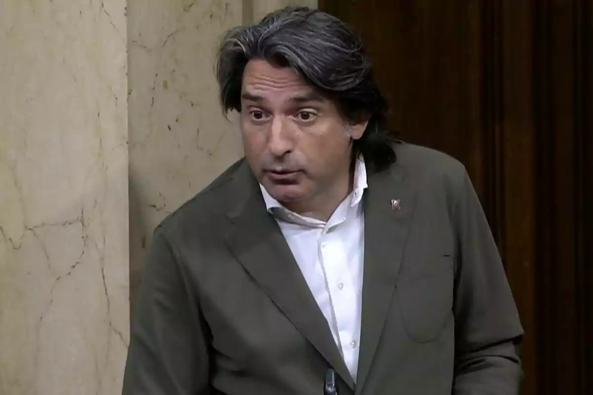 Hombre de cabello canoso y largo con saco verde oliva y camisa blanca hablando en un entorno formal con paredes de mármol y madera.