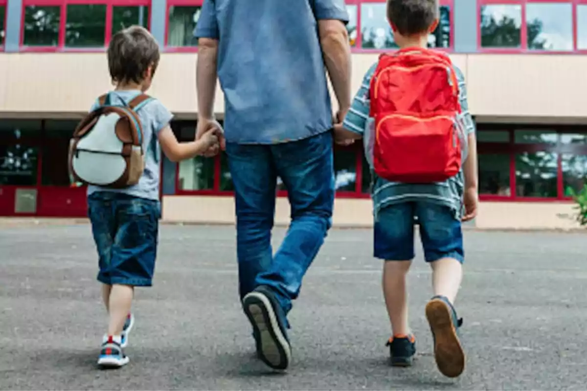 Persona adulta caminando de la mano con dos niños pequeños que llevan mochilas frente a un edificio escolar