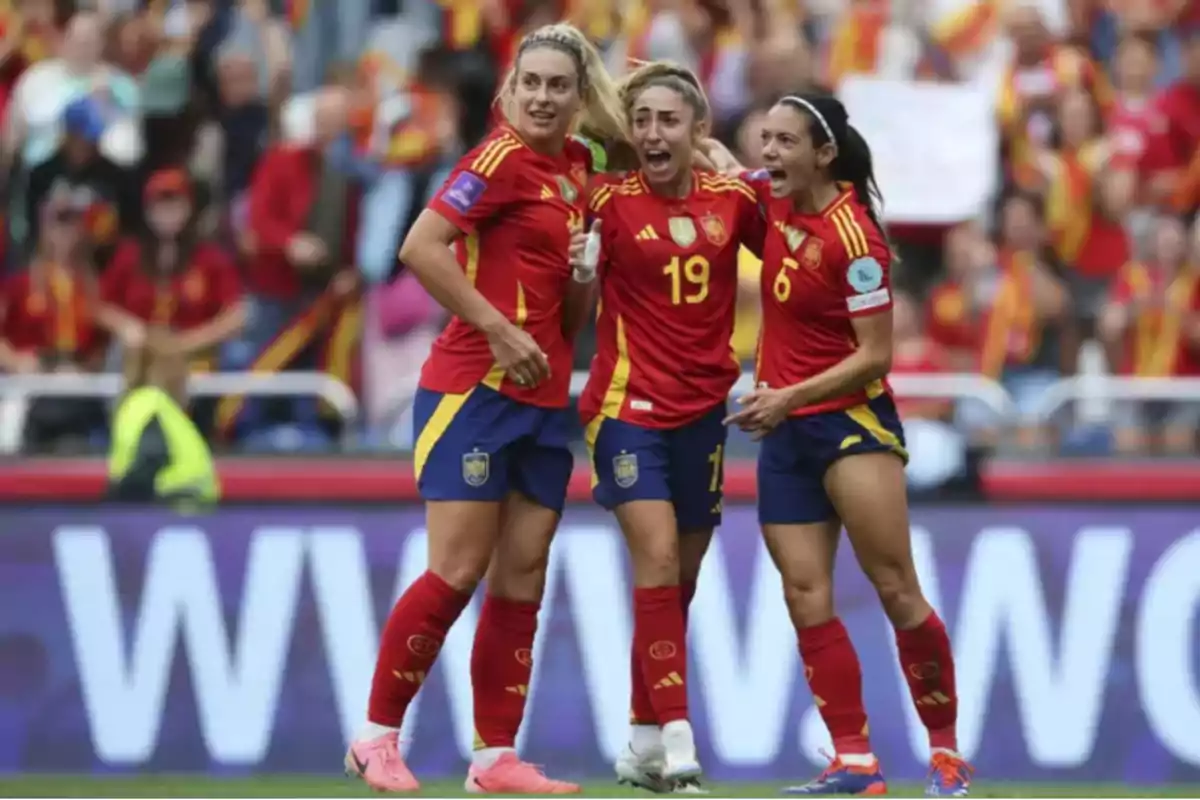 Tres jugadoras de la selección femenina de fútbol de España celebran un gol en el campo con expresiones de alegría y emoción mientras el público las observa desde las gradas Tres jugadoras de la selección femenina de fútbol de España celebran un gol en el campo con expresiones de alegría y emoción mientras el público las observa desde las gradas
