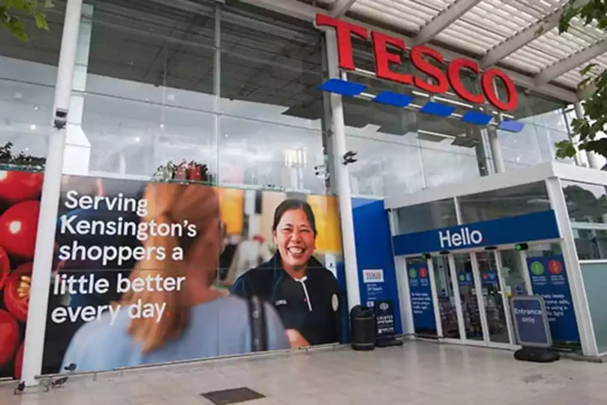 Fachada de una tienda Tesco con un gran cartel publicitario que muestra a una empleada sonriendo y la entrada principal con puertas automáticas Fachada de una tienda Tesco con un gran cartel publicitario que muestra a una empleada sonriendo y la entrada principal con puertas automáticas