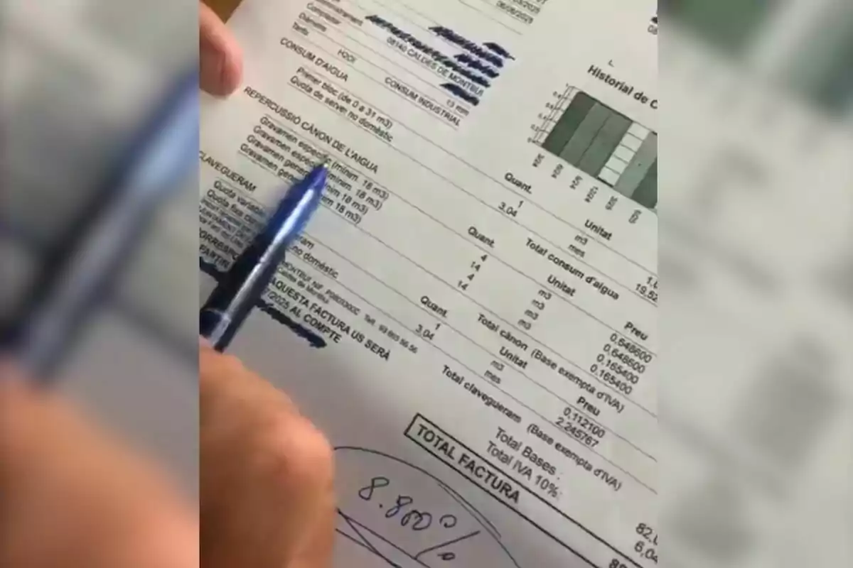 Persona señalando con una pluma azul una factura de agua escrita en catalán donde se observan datos de consumo, tarifas y un gráfico de historial de consumo en la parte superior derecha