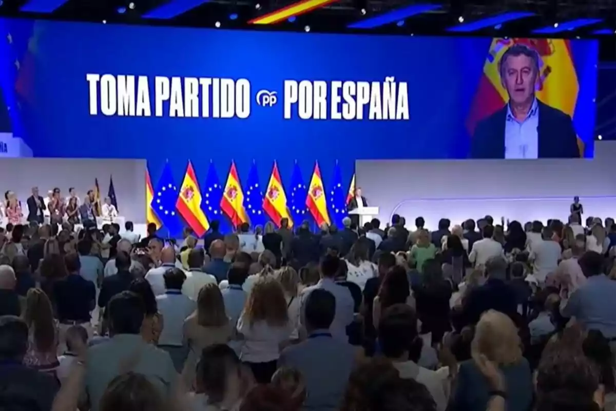 Auditorio lleno de personas asistiendo a un evento político del Partido Popular en España con banderas españolas y europeas en el escenario y una gran pantalla con el mensaje Toma partido por España Auditorio lleno de personas asistiendo a un evento político del Partido Popular en España con banderas españolas y europeas en el escenario y una gran pantalla con el mensaje Toma partido por España