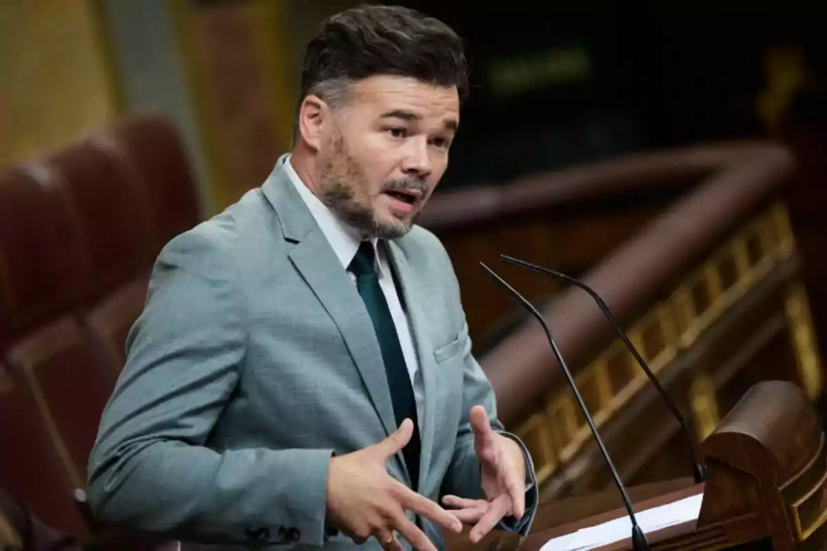 Hombre con barba y cabello oscuro vestido con traje gris y corbata oscura hablando en un podio en un recinto parlamentario Hombre con barba y cabello oscuro vestido con traje gris y corbata oscura hablando en un podio en un recinto parlamentario