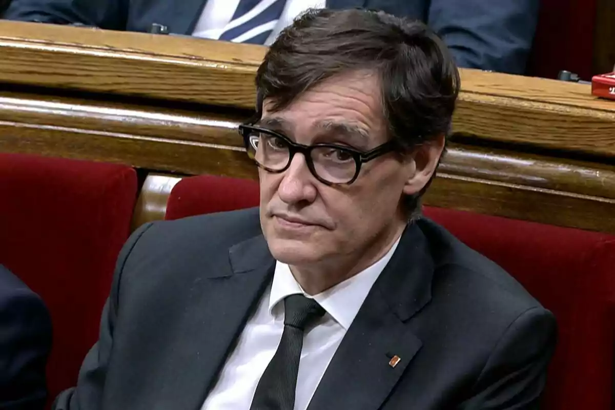 Hombre con gafas y traje oscuro sentado en un banco rojo en un entorno formal