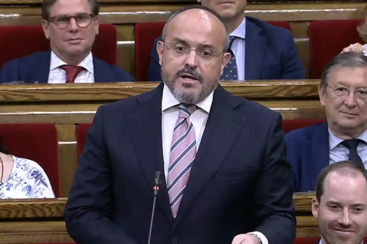 Hombre con barba y gafas hablando en un parlamento mientras otras personas lo observan desde sus asientos Hombre con barba y gafas hablando en un parlamento mientras otras personas lo observan desde sus asientos