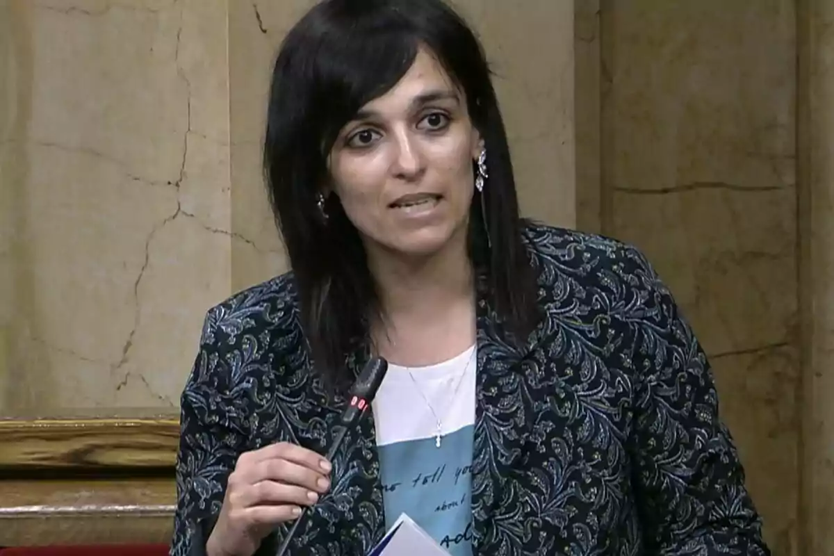 Mujer de cabello oscuro hablando frente a un micrófono en un entorno formal con paredes de mármol.