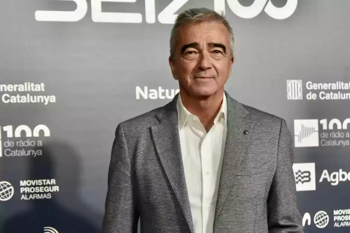 Hombre de cabello canoso con saco gris y camisa blanca posando frente a un fondo con logotipos y texto en un evento Hombre de cabello canoso con saco gris y camisa blanca posando frente a un fondo con logotipos y texto en un evento