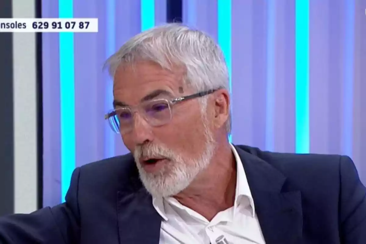 David Cantero de cabello y barba canosos con gafas y traje oscuro hablando en un programa de televisión con fondo azul David Cantero de cabello y barba canosos con gafas y traje oscuro hablando en un programa de televisión con fondo azul