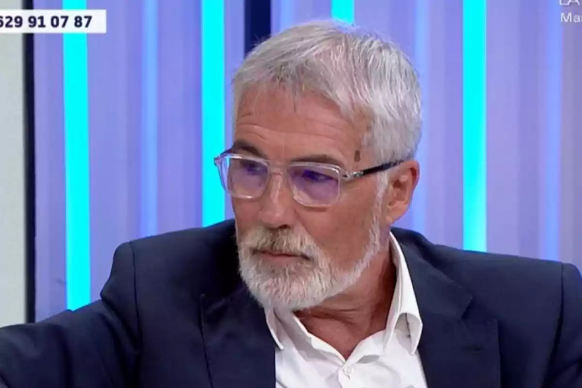 David Cantero de cabello y barba canosos con gafas transparentes y traje oscuro sentado en un estudio de televisión con luces azules de fondo David Cantero de cabello y barba canosos con gafas transparentes y traje oscuro sentado en un estudio de televisión con luces azules de fondo