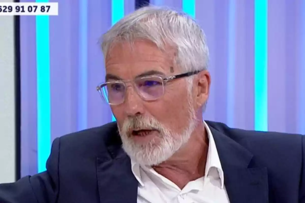 David Cantero de cabello y barba canosos con gafas transparentes y saco oscuro hablando en un set de televisión con luces azules de fondo David Cantero de cabello y barba canosos con gafas transparentes y saco oscuro hablando en un set de televisión con luces azules de fondo