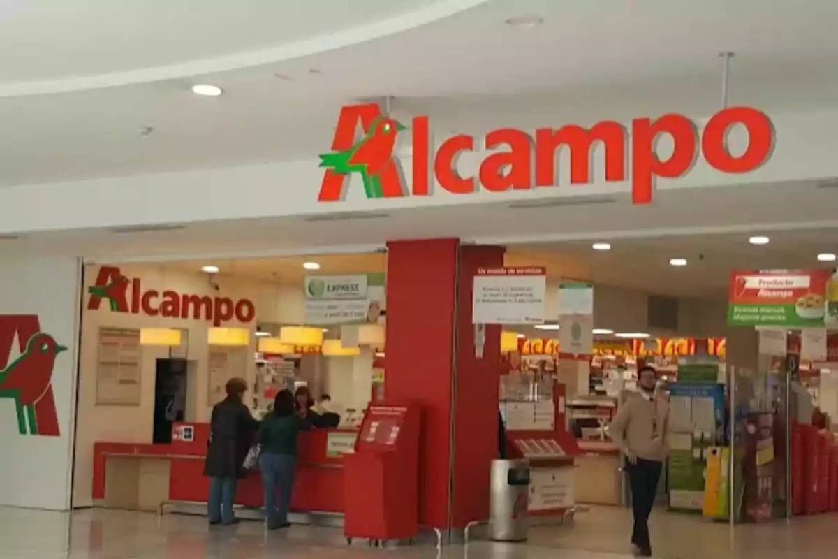 Entrada de un supermercado Alcampo con personas en la zona de atención al cliente y el logo visible en la parte superior. Entrada de un supermercado Alcampo con personas en la zona de atención al cliente y el logo visible en la parte superior.
