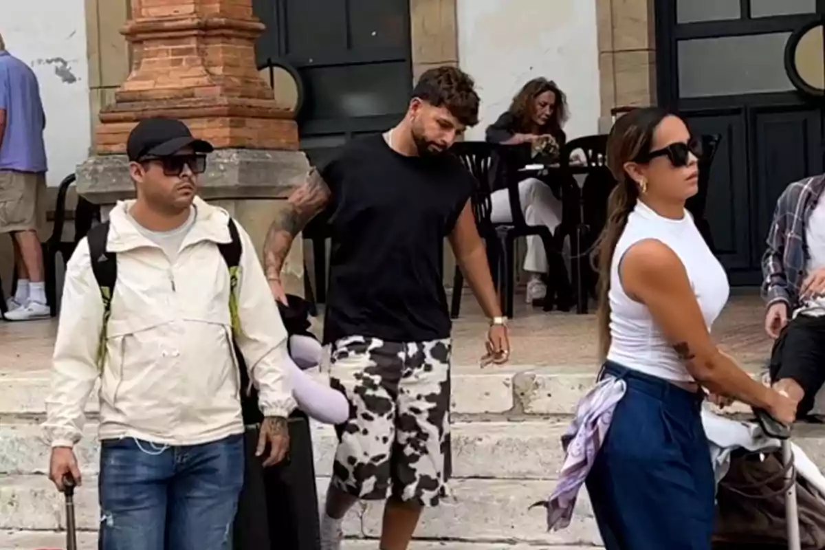 Gloria Camila y José Fernando caminan por la calle con maletas mientras una mujer está sentada al fondo en una mesa Gloria Camila y José Fernando caminan por la calle con maletas mientras una mujer está sentada al fondo en una mesa
