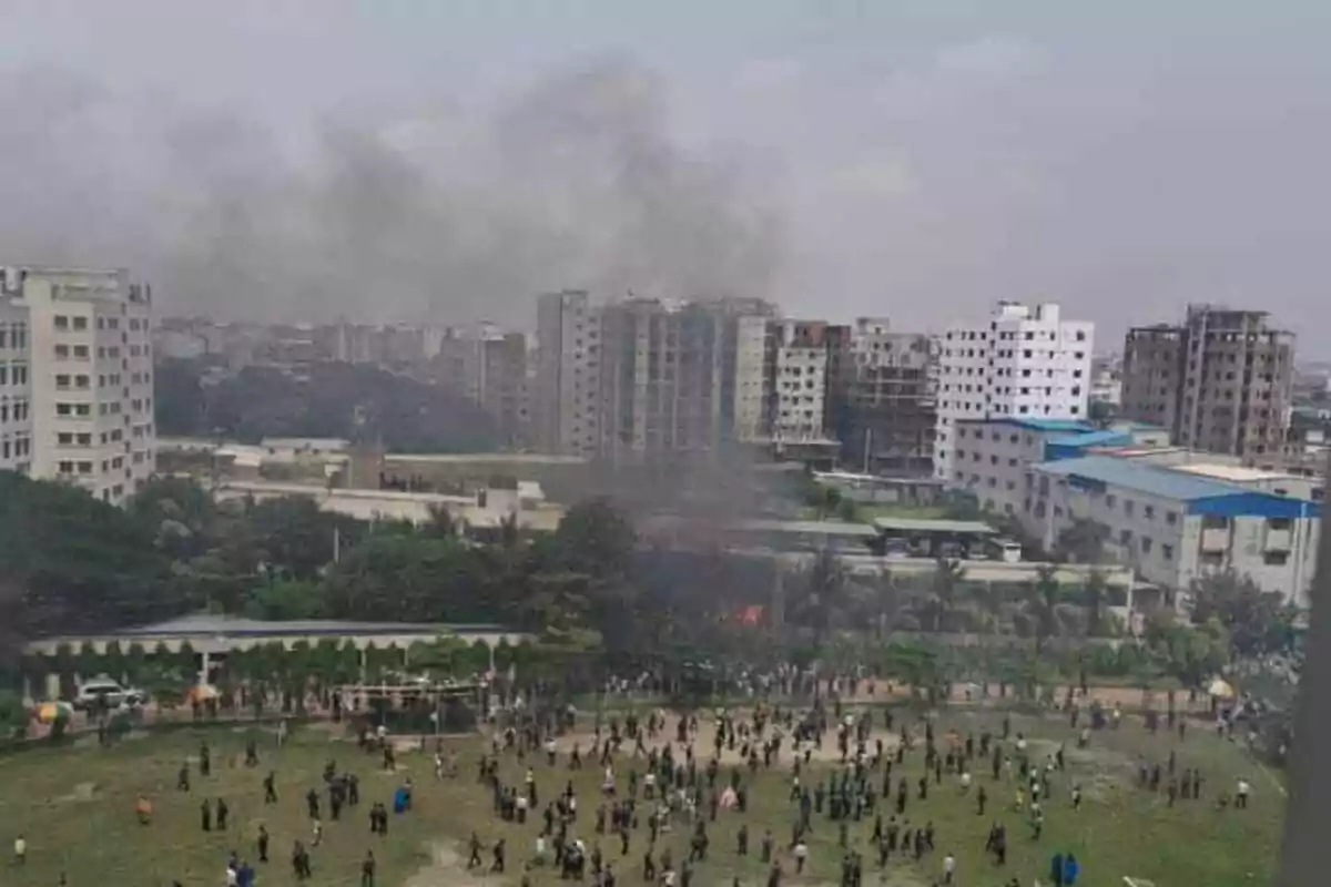 Personas reunidas en un campo mientras una columna de humo se eleva desde un área con edificios al fondo en una ciudad Personas reunidas en un campo mientras una columna de humo se eleva desde un área con edificios al fondo en una ciudad