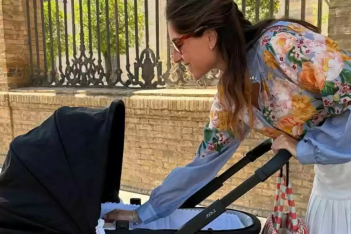 Lourdes Montes vestida con una blusa de flores y gafas de sol mirando y tocando un cochecito de bebé negro en la calle. Lourdes Montes vestida con una blusa de flores y gafas de sol mirando y tocando un cochecito de bebé negro en la calle.