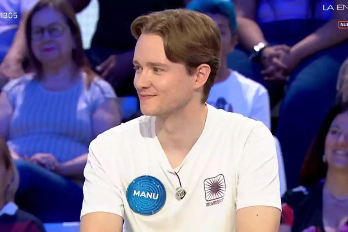 Manu con camiseta blanca y placa azul que dice Manu participando en un programa de televisión con público de fondo Manu con camiseta blanca y placa azul que dice Manu participando en un programa de televisión con público de fondo