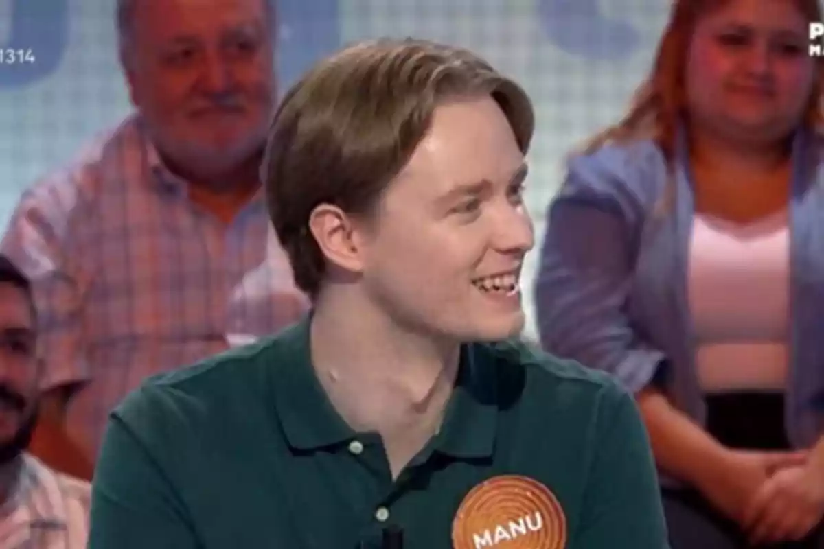 Manu sonriente con una camiseta verde y una chapa que dice Manu participa en un programa de televisión con público al fondo Manu sonriente con una camiseta verde y una chapa que dice Manu participa en un programa de televisión con público al fondo