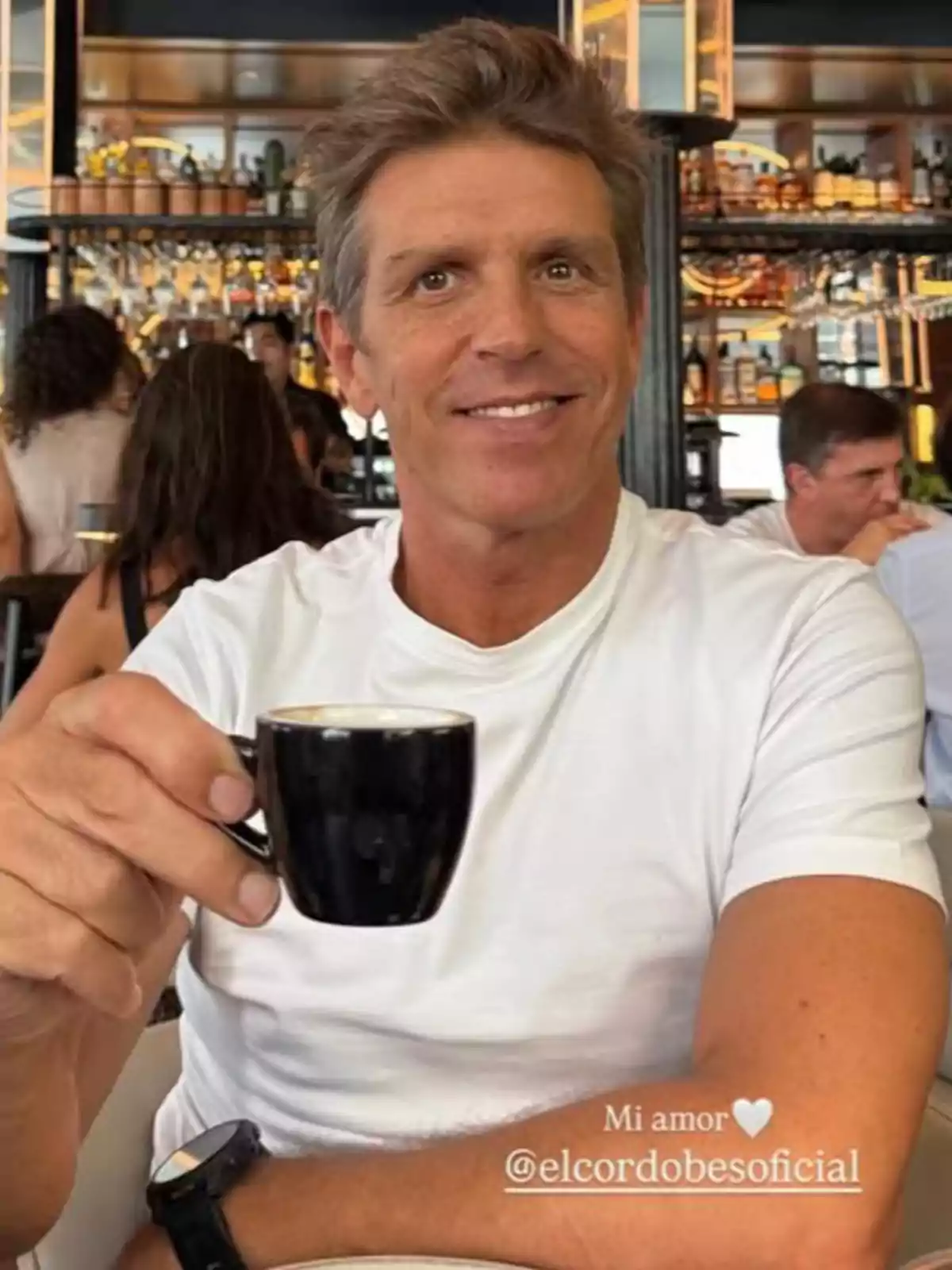 Manuel Díaz ‘El Cordobés’ sonriente con camiseta blanca sosteniendo una taza de café en una cafetería elegante Manuel Díaz ‘El Cordobés’ sonriente con camiseta blanca sosteniendo una taza de café en una cafetería elegante