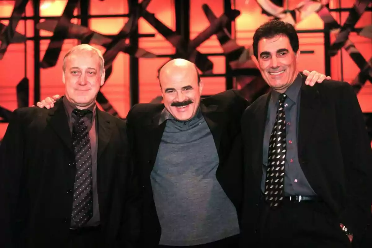 Tres hombres vestidos de traje sonríen y posan juntos frente a un fondo iluminado de color naranja Tres hombres vestidos de traje sonríen y posan juntos frente a un fondo iluminado de color naranja