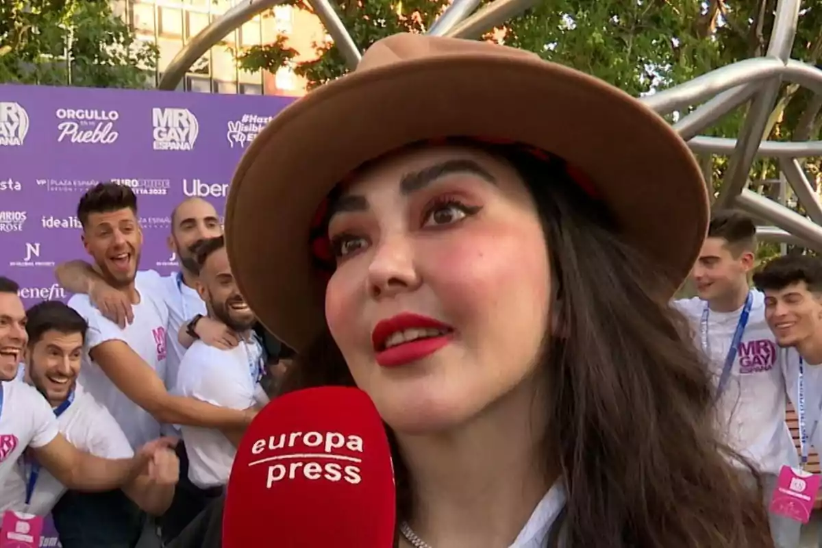 Patricia Donoso con sombrero y maquillaje llamativo es entrevistada en un evento al aire libre con un grupo de personas sonrientes al fondo y un micrófono rojo de europa press en primer plano Patricia Donoso con sombrero y maquillaje llamativo es entrevistada en un evento al aire libre con un grupo de personas sonrientes al fondo y un micrófono rojo de europa press en primer plano
