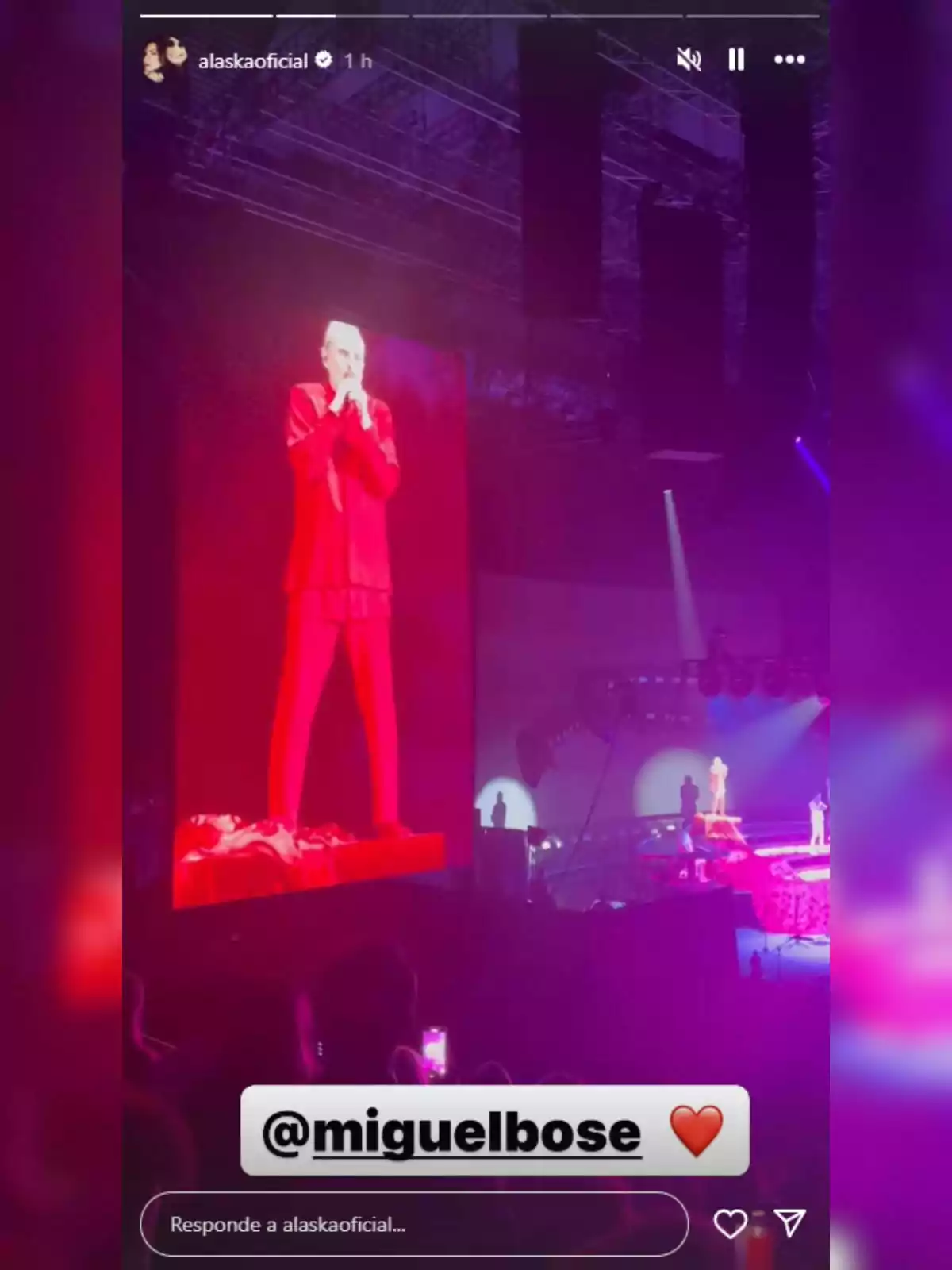 Miguel Bosé vestido de rojo se presenta en un escenario iluminado, mientras su imagen aparece en una pantalla gigante y el público observa el espectáculo. Miguel Bosé vestido de rojo se presenta en un escenario iluminado, mientras su imagen aparece en una pantalla gigante y el público observa el espectáculo.