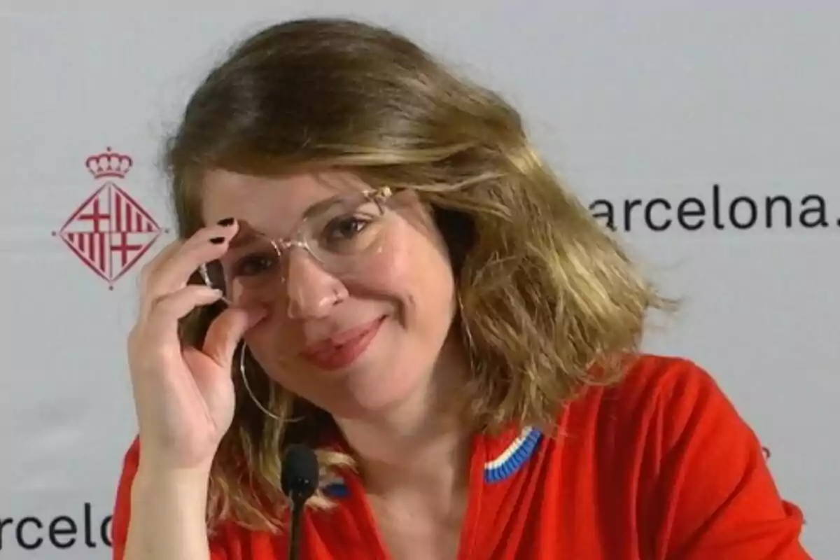 Mujer con gafas y cabello castaño sonríe mientras sostiene sus lentes con una mano, viste una camisa roja y está frente a un micrófono con un fondo que tiene el logo de Barcelona
