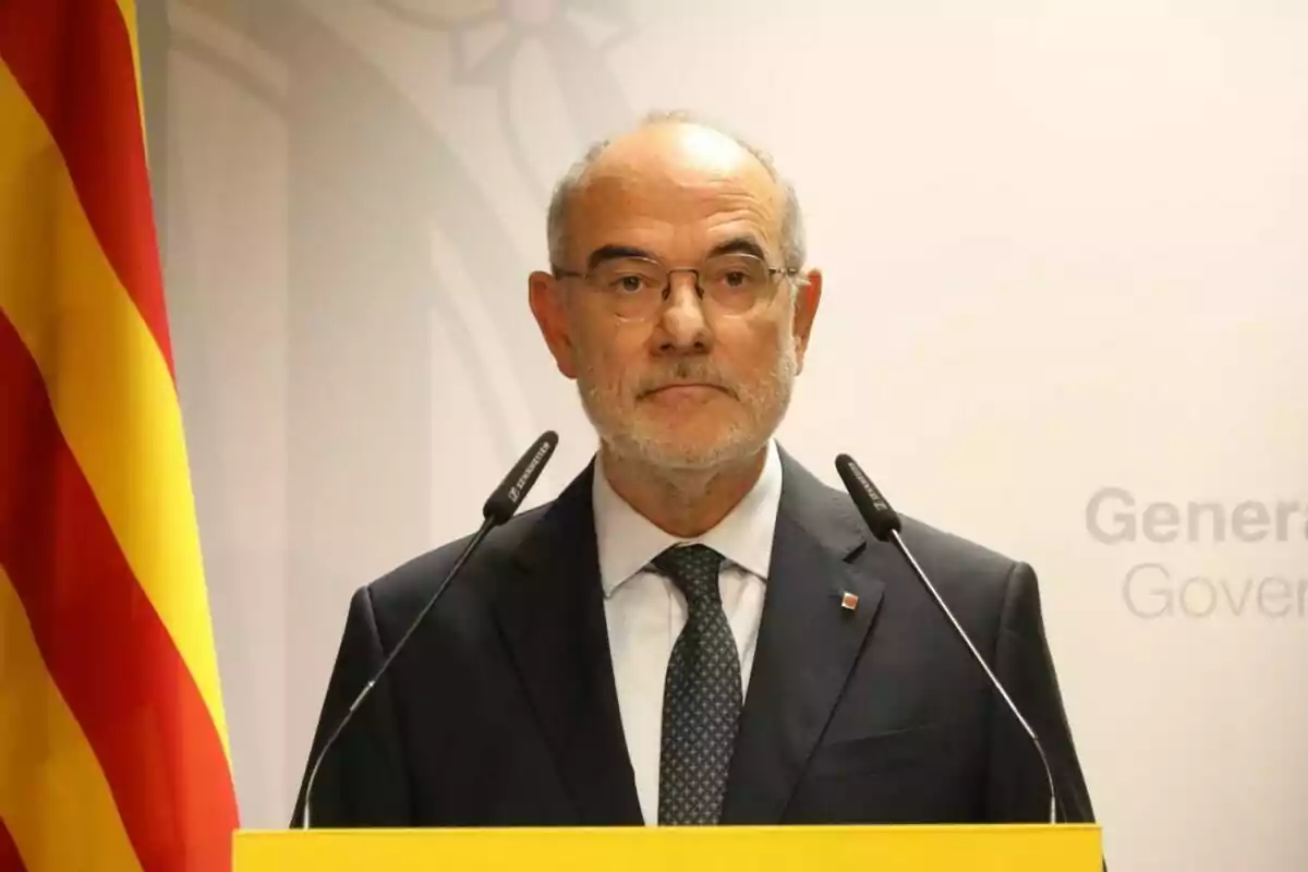 Hombre mayor con gafas y barba canosa hablando en una rueda de prensa con micrófonos y una bandera catalana a su lado Hombre mayor con gafas y barba canosa hablando en una rueda de prensa con micrófonos y una bandera catalana a su lado