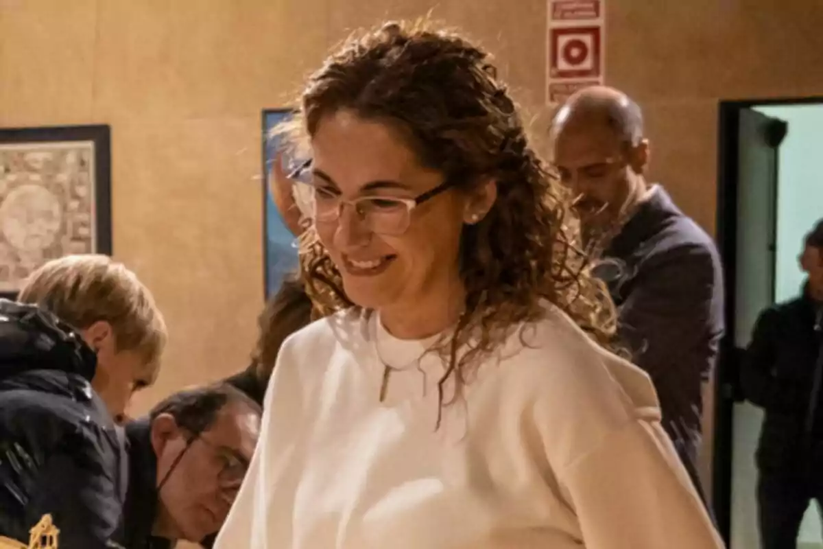 Mujer de cabello rizado y gafas sonríe mientras participa en una actividad en un espacio interior con varias personas al fondo Mujer de cabello rizado y gafas sonríe mientras participa en una actividad en un espacio interior con varias personas al fondo