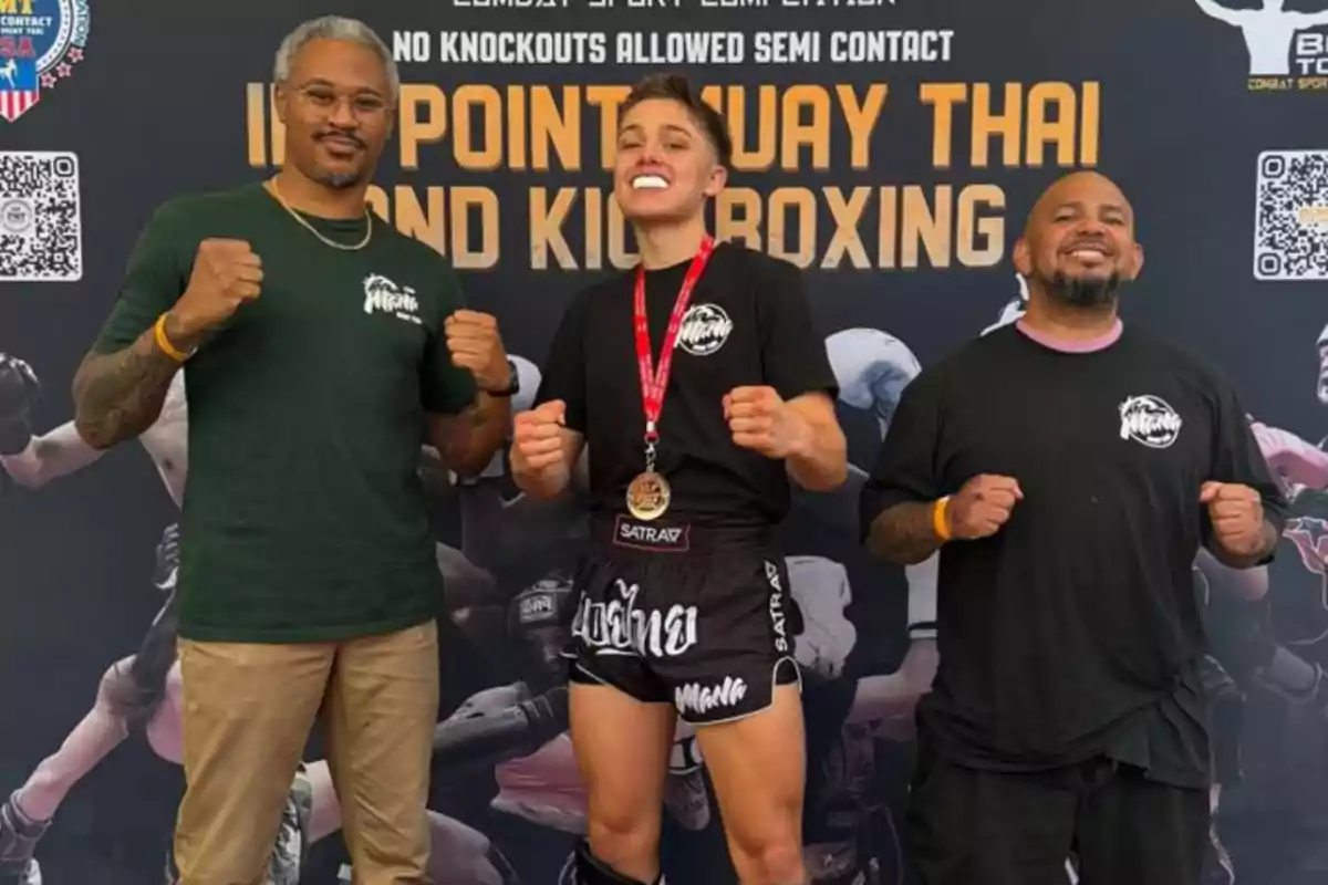 Knox Jolie Pitt junto al dos coach posan sonrientes con actitud de victoria frente a un cartel de Muay Thai y Kickboxing, la persona del centro lleva una medalla y protector bucal Knox Jolie Pitt junto al dos coach posan sonrientes con actitud de victoria frente a un cartel de Muay Thai y Kickboxing, la persona del centro lleva una medalla y protector bucal