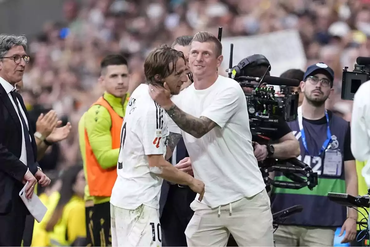 Un futbolista con uniforme blanco es abrazado por un hombre vestido de blanco mientras varias personas observan y una cámara profesional graba la escena en un estadio lleno de público. Un futbolista con uniforme blanco es abrazado por un hombre vestido de blanco mientras varias personas observan y una cámara profesional graba la escena en un estadio lleno de público.