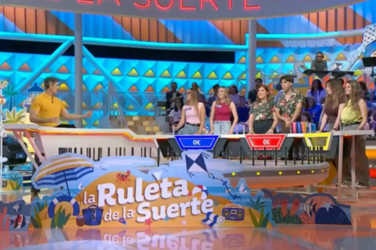Un grupo de concursantes y Jorge Fernández participan en el set colorido del programa de televisión La Ruleta de la Suerte con decoración veraniega y público al fondo. Captura de 'La Ruleta de la Suerte' Un grupo de concursantes y Jorge Fernández participan en el set colorido del programa de televisión La Ruleta de la Suerte con decoración veraniega y público al fondo. Captura de 'La Ruleta de la Suerte'