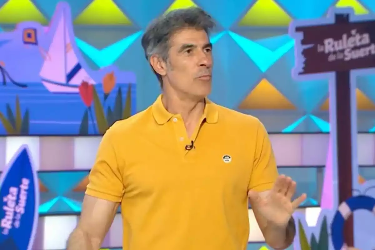 Jorge Fernández con camiseta amarilla en un set colorido de televisión con un letrero de La Ruleta de la Suerte al fondo Jorge Fernández con camiseta amarilla en un set colorido de televisión con un letrero de La Ruleta de la Suerte al fondo