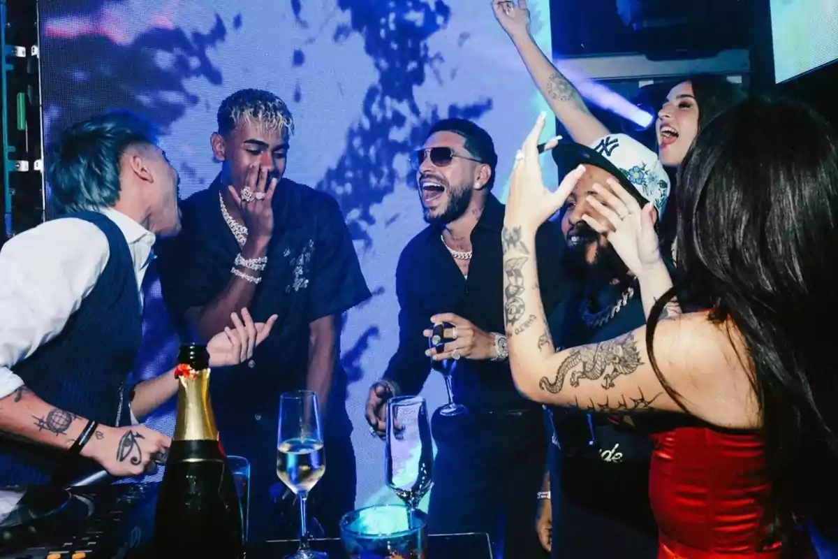 Un grupo de personas jóvenes disfruta de una fiesta en un club nocturno, riendo y celebrando con copas y botellas sobre la mesa, rodeados de luces azules y ambiente animado. Un grupo de personas jóvenes disfruta de una fiesta en un club nocturno, riendo y celebrando con copas y botellas sobre la mesa, rodeados de luces azules y ambiente animado.