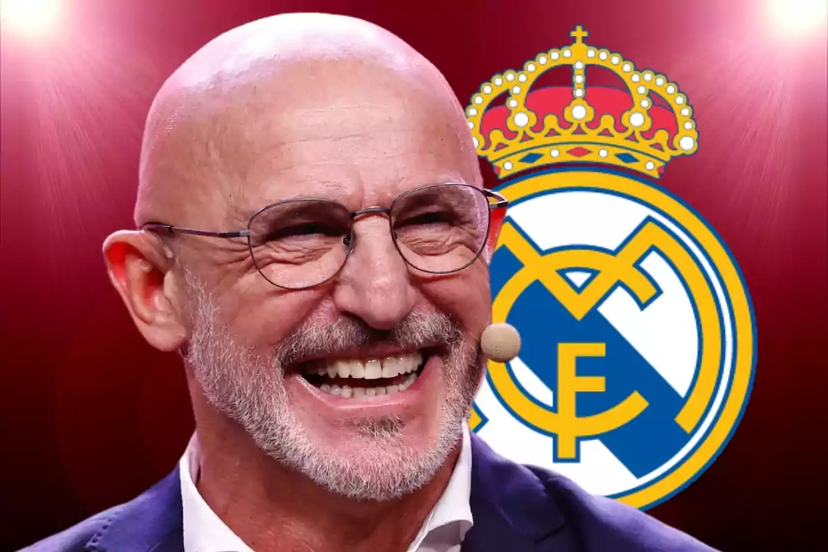 Hombre calvo con gafas y micrófono sonríe frente al escudo del Real Madrid sobre un fondo rojo