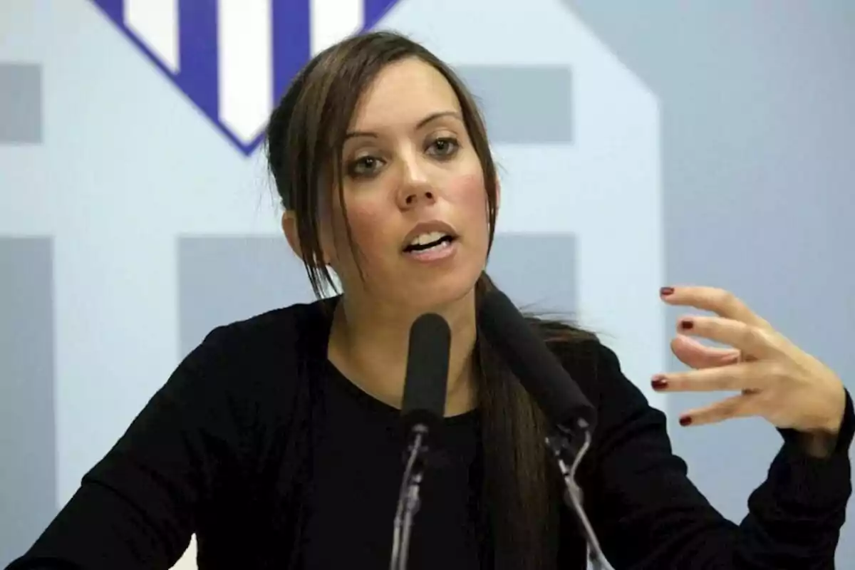 Mujer joven hablando frente a dos micrófonos con fondo azul claro y un logo geométrico detrás