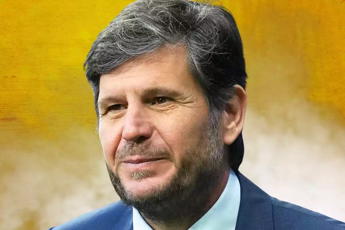 Hombre de cabello canoso y barba corta vistiendo saco azul con fondo amarillo difuminado